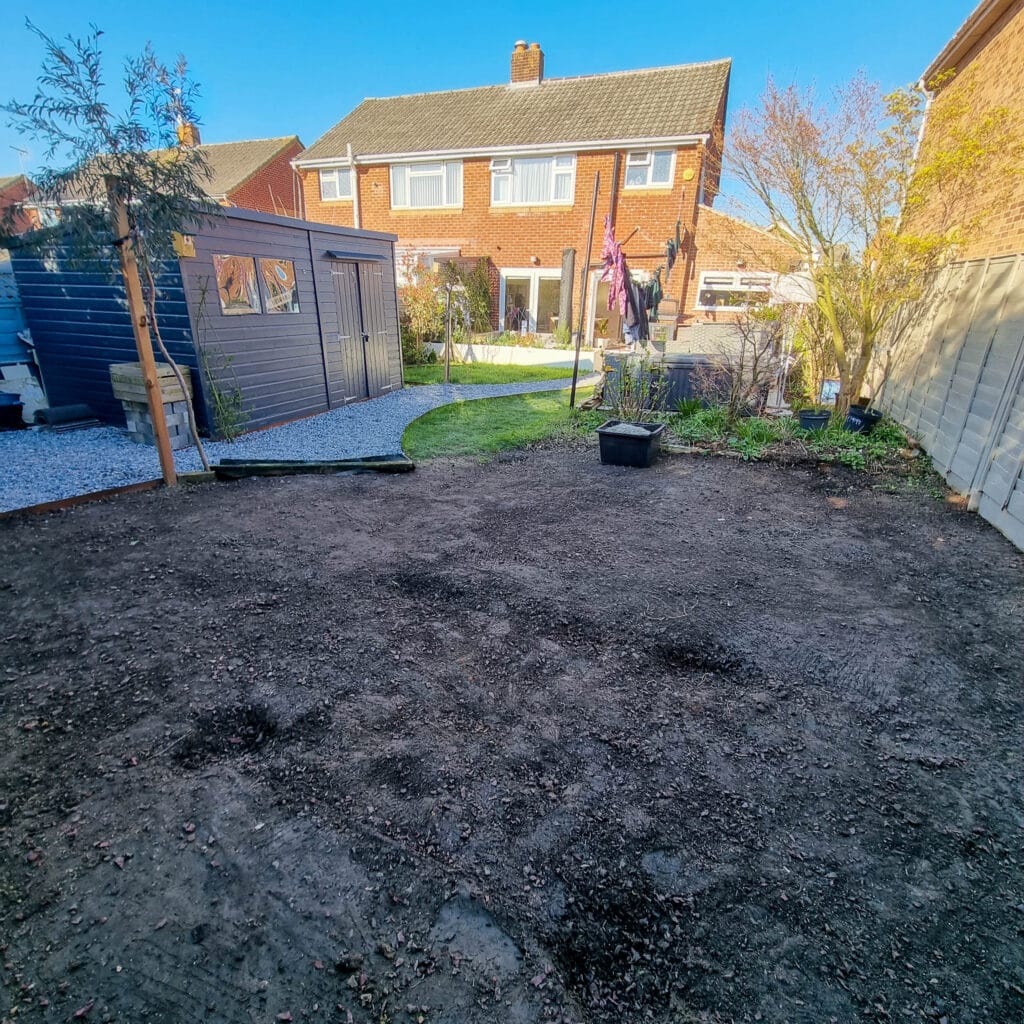A back garden with a patch of bare soil in the foreground, a small shed on the left, a lawn, potted plants, and space for a DIY pergola. A brick house and garden are visible in the background under a clear blue sky.