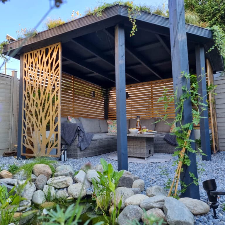 A stylish outdoor gazebo with a green roof, wooden slatted walls, and a decorative panel creates the perfect DIY pergola. Inside are grey cushioned chairs, snacks and blankets. The foreground features a small pond for a true wildlife garden feel.