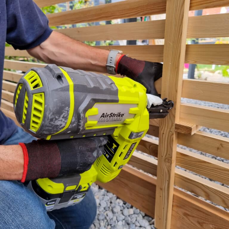 A person wearing gloves uses a yellow cordless nail gun to assemble a wooden slat fence outdoors in a wildlife garden, with gravel visible on the ground.