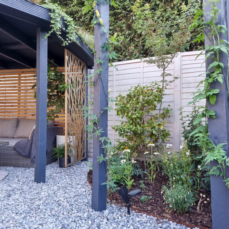 A modern wildlife garden with pea gravel, dark wooden posts with climbing plants, a cosy seating area under a DIY pergola, a decorative wooden screen, and lush greenery along a light-coloured fence.