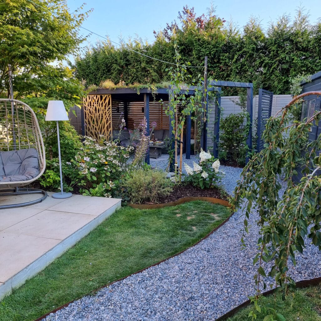 A landscaped wildlife garden with a gravel path, green lawn, hanging chair, and a stylish DIY pergola seating area surrounded by lush plants and trees on a sunny day.
