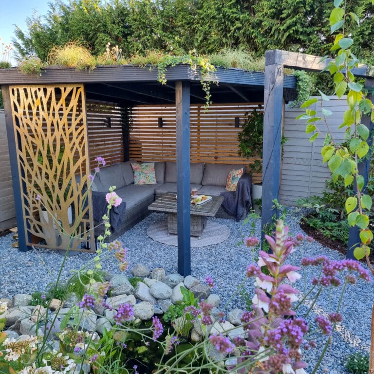 A cosy outdoor seating area with grey sofas under a DIY pergola, surrounded by lush plants, flowers, and a small rock garden. A gold decorative screen adds style, making this space a relaxing garden retreat in our latest Garden Reveal.