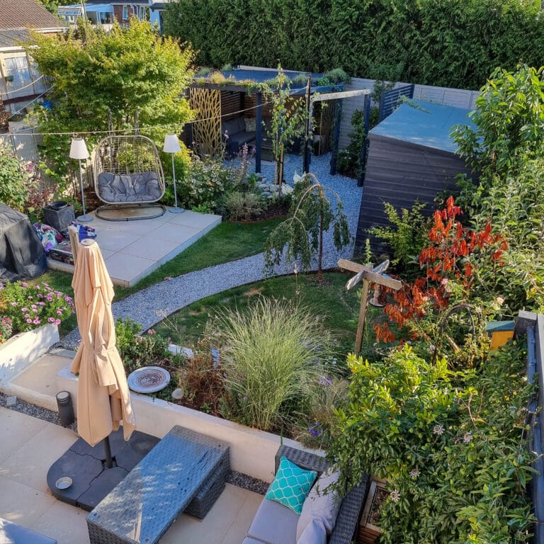 A well-maintained garden features a patio with seating, a parasol, lush plants, a small lawn, a gravel path, a swing seat, DIY pergola, shed, and colourful bushes and trees—all bordered by tall hedges.