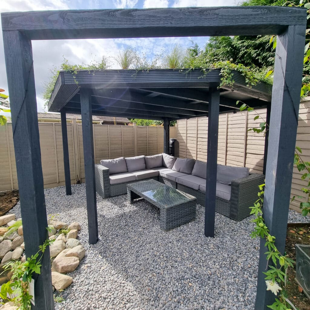 A modern outdoor seating area with a grey wicker sectional sofa and a glass-topped coffee table sits under a black pergola on gravel, surrounded by greenery and wooden privacy fencing enhanced with stylish batten screening.