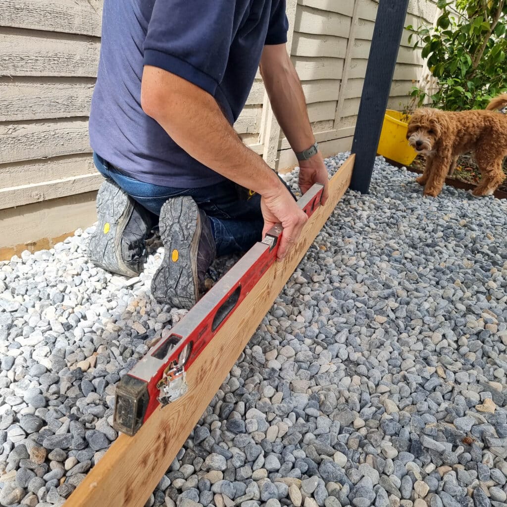 A person kneels on gravel using a spirit level to measure a wooden plank for batten screening outdoors. A small brown dog stands nearby, and a wooden fence is in the background.