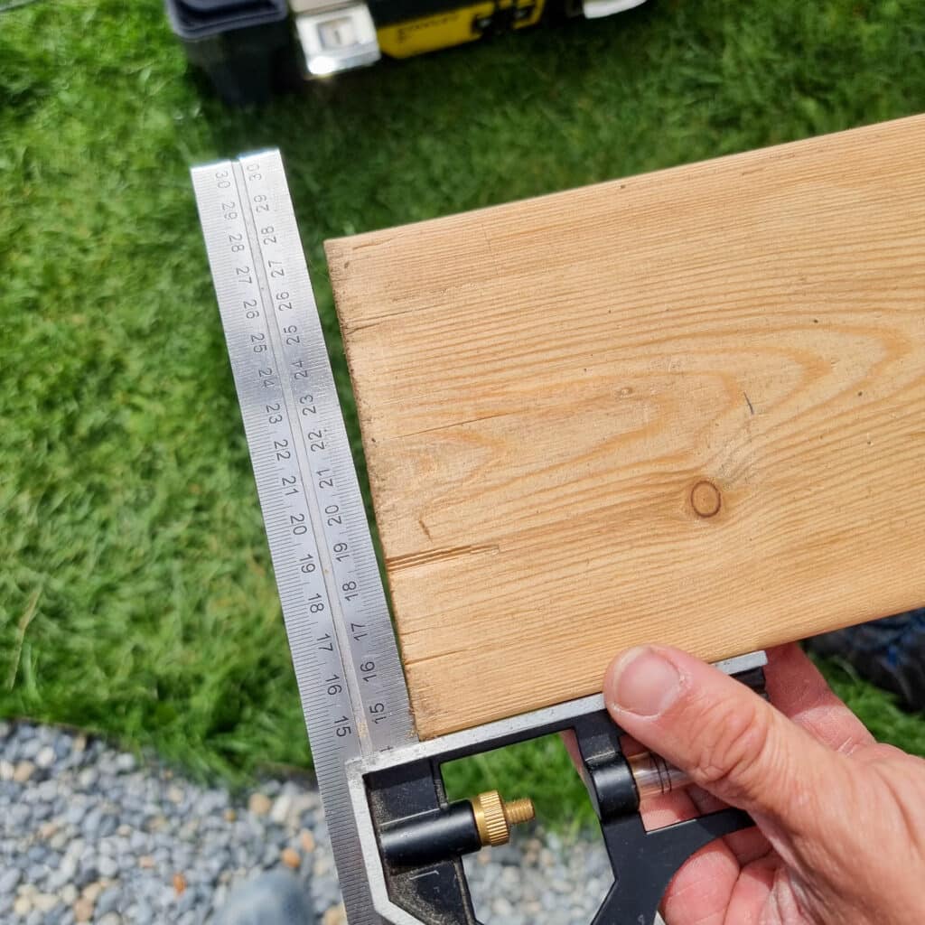 A hand holds a carpenter’s square against the edge of a wooden board outdoors, measuring and checking the angle for batten screening, with grass and gravel visible in the background.