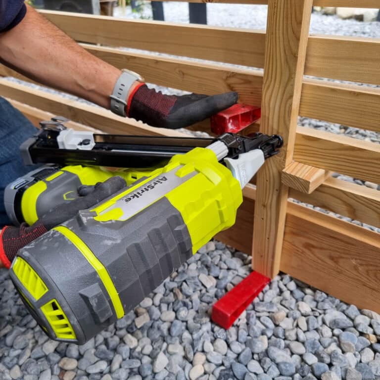 A person uses a green and grey Ryobi nail gun to attach wooden slats for batten screening to a vertical post, holding the wood in place with red spacers. The person wears gloves and works on a pebble-covered ground.