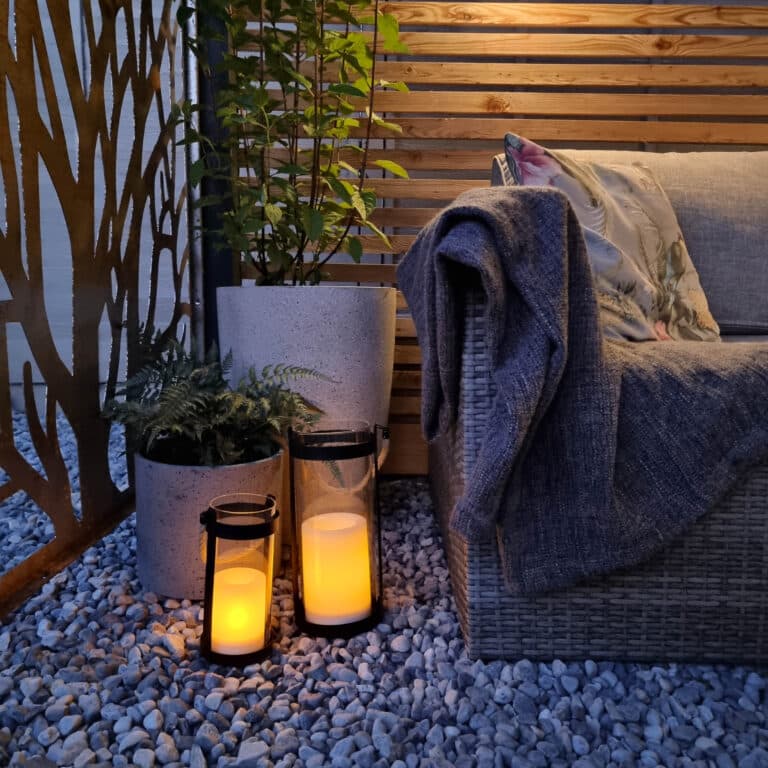 Cosy outdoor seating area with a wicker sofa, floral cushion, and grey blanket. Two lanterns with solar powered lighting and two potted plants sit on pebble stones, framed by timber panel fencing in the background.