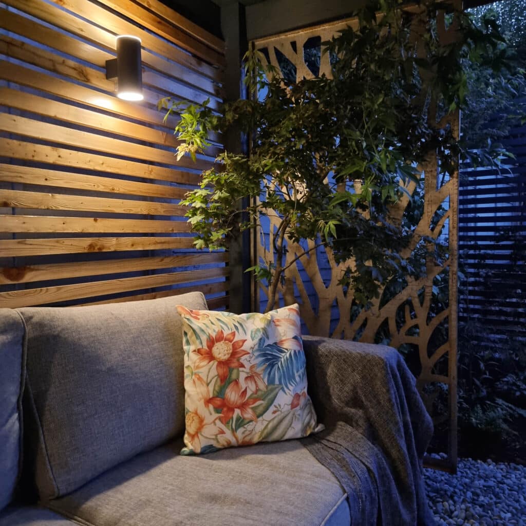 A cosy outdoor patio at dusk features a grey sofa with a floral cushion and blanket, soft solar lighting, wooden slats, leafy plants, and decorative privacy screens beneath a charming pergola in the background.