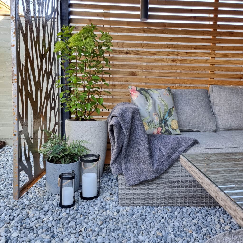 A modern outdoor patio with a grey wicker sofa, floral cushion, grey throw blanket, potted plants, solar powered lighting in two lanterns with white candles, and a wooden slat privacy screen on gravel flooring.