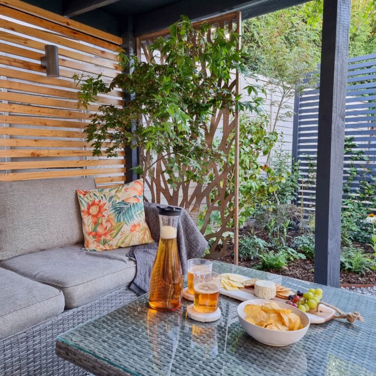 A cosy patio with a grey wicker sofa, a floral cushion, and a glass table set with iced tea, cheese and fruit platter, and crisps. Green plants and decorative screens surround the space beneath a pergola with solar powered lighting.