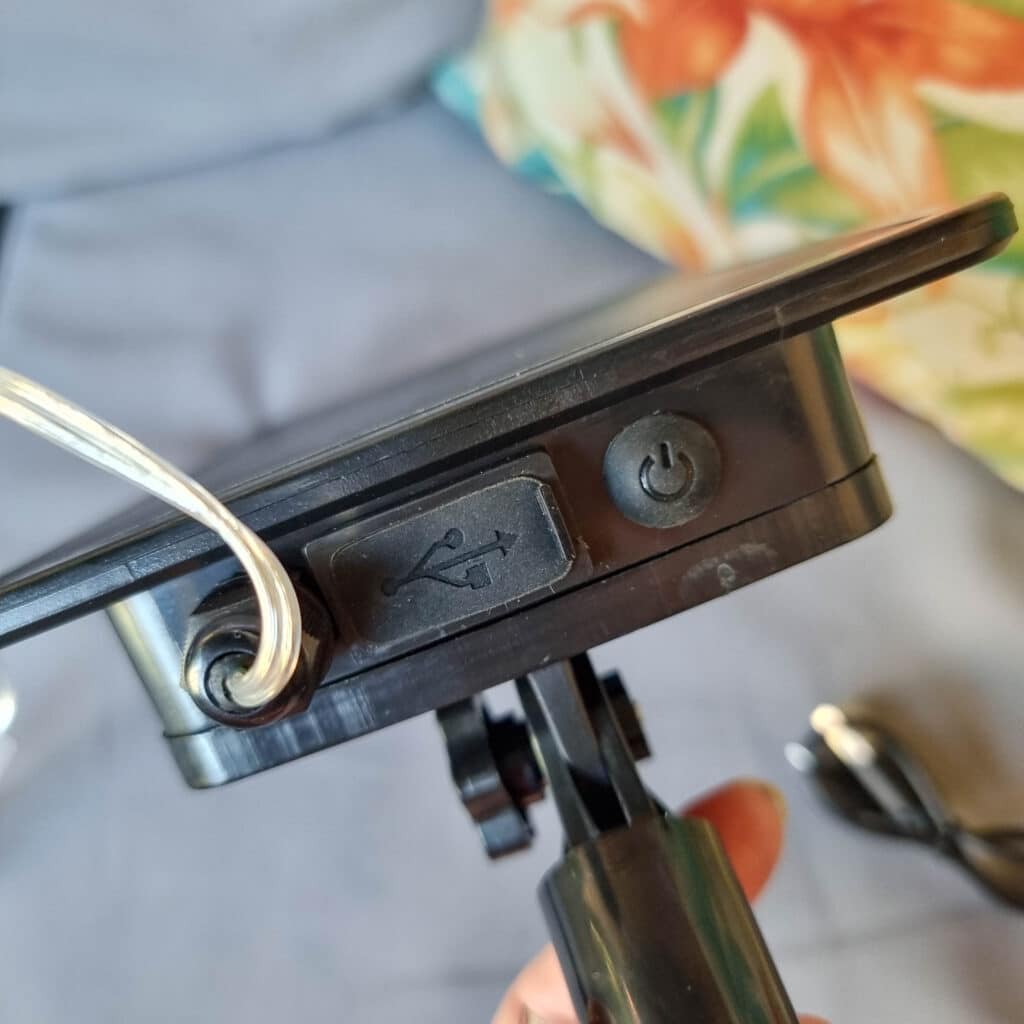 Close-up of a black device with a USB cable plugged in, a USB port symbol, and a power button—ideal for solar powered lighting setups. The background features a floral-patterned cushion and a grey surface.