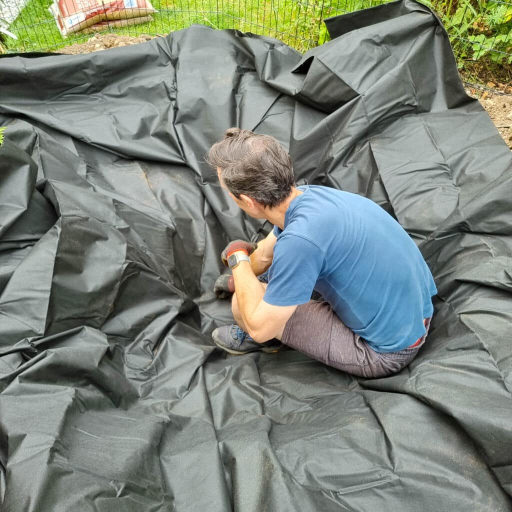 A man kneels on a large black landscape fabric, adjusting it as he works outdoors in a garden area, preparing for wildlife pond construction. Soil, grass, and gardening supplies are visible around the fabric.
