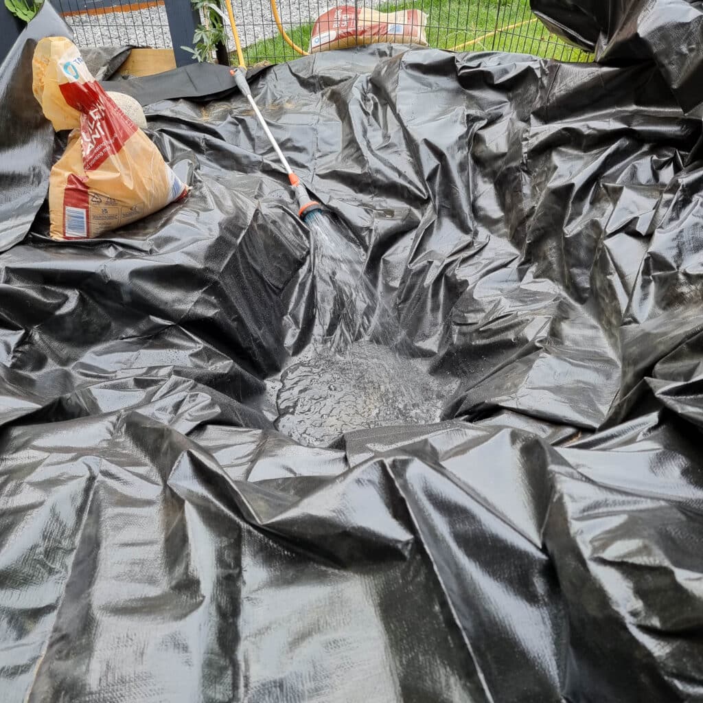 A black pond liner is being fitted into a shallow hole in the ground as part of wildlife pond construction. A bag of sand and a garden tool are nearby, with water pooling in the centre. Grass and a fence can be seen in the background.