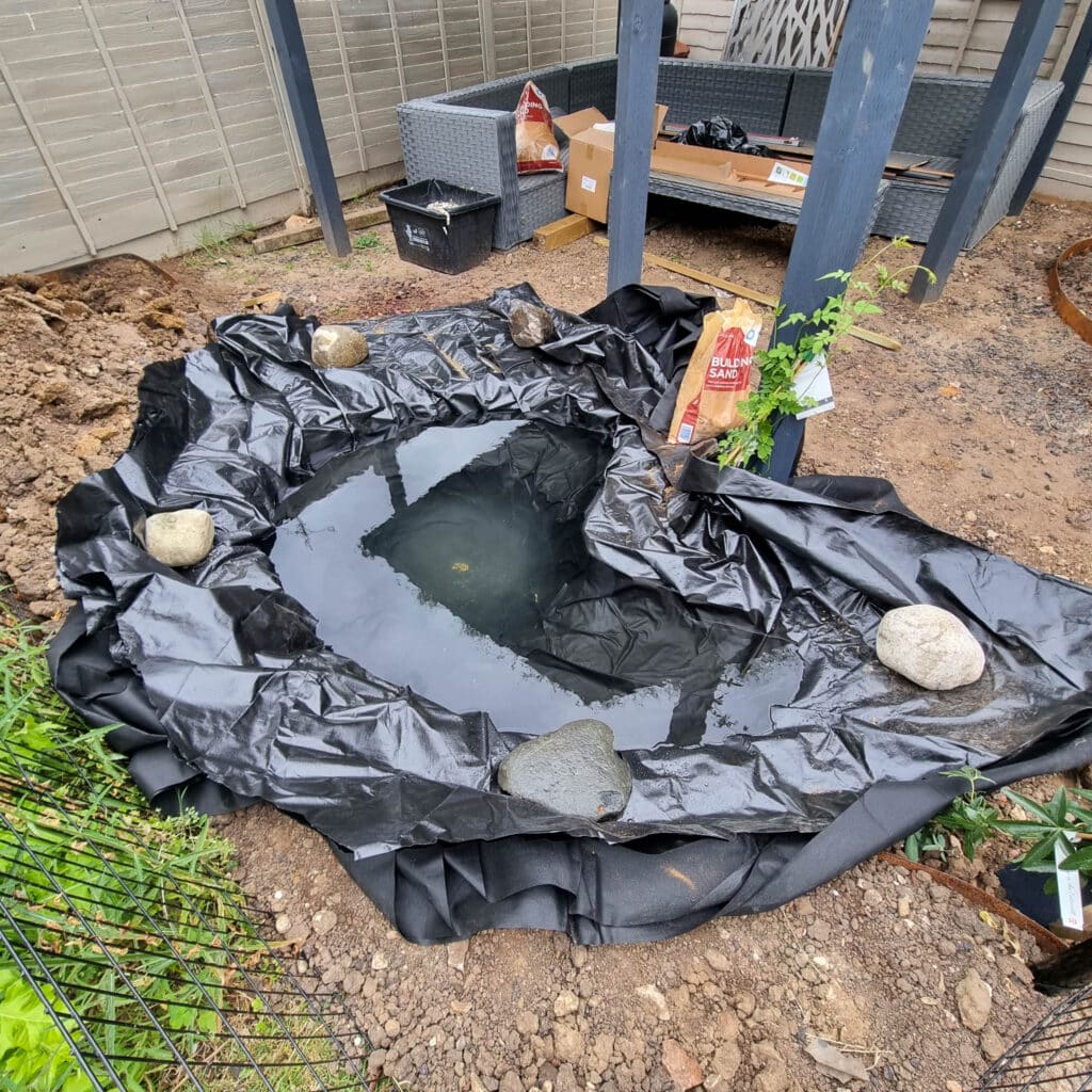 A back garden wildlife pond under construction with a black liner secured by large rocks and partially filled with water. Gardening materials, soil, and patio furniture are visible in the background, enhancing this future wildlife garden.