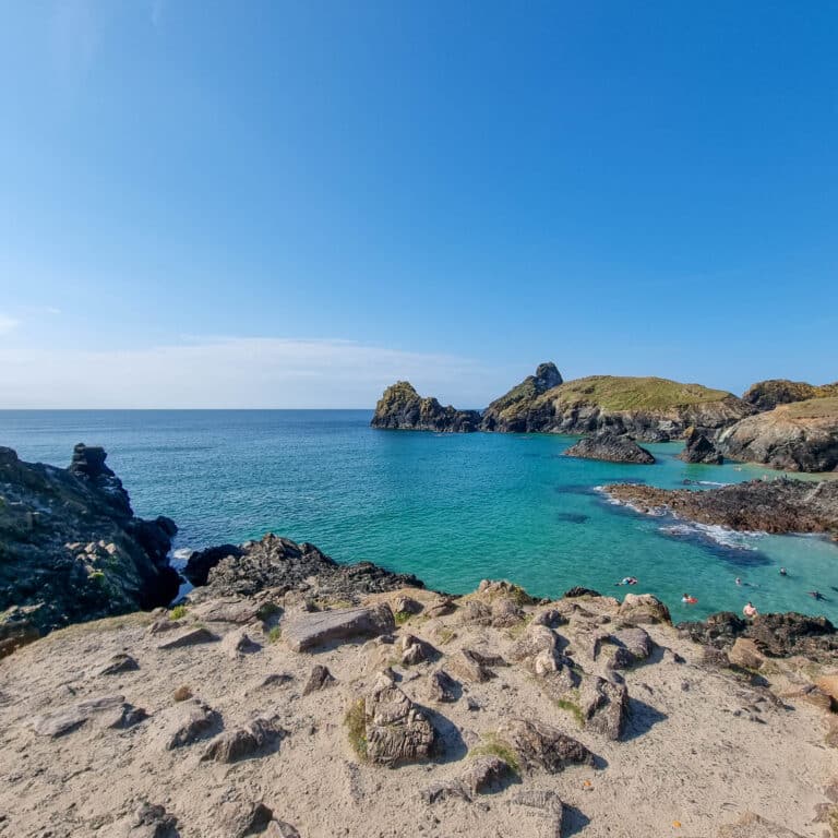 Rocky shoreline with turquoise water and swimmers in a small cove, bordered by rugged cliffs and grassy hills under a clear blue sky—a serene escape perfect for the best holidays or peaceful addiction recovery.