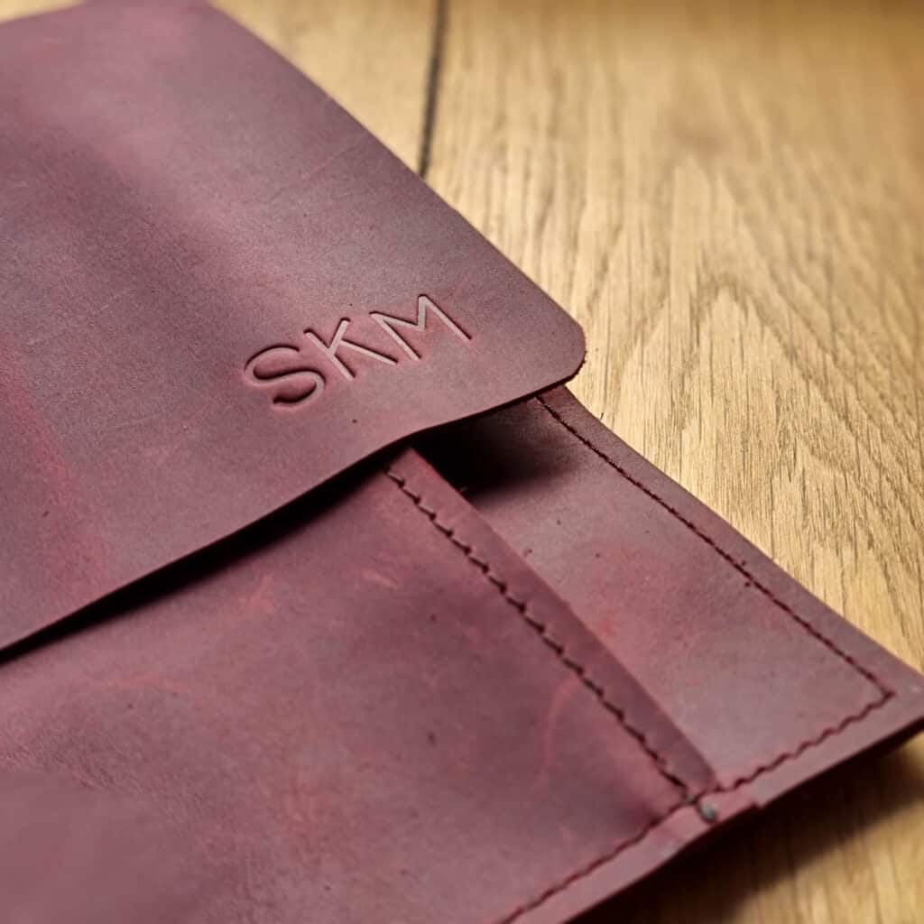 A close-up of a brown leather pouch with visible stitching and the initials SKM embossed on the flap, placed on a wooden surface—an elegant accessory to enhance your work from home environment.