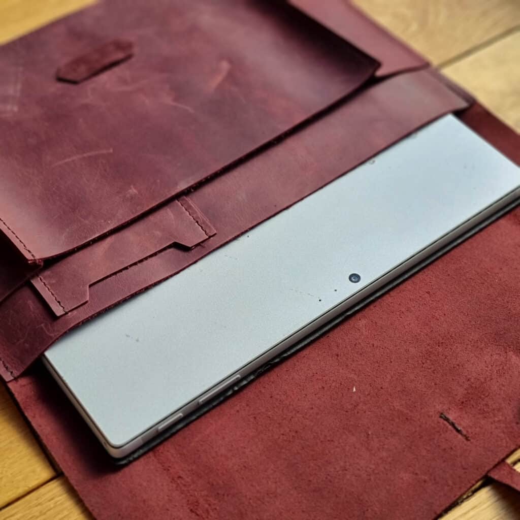A silver laptop partially slid into a brown leather sleeve cover, lying on a wooden surface—perfect for creating a good working environment as you work from home. The sleeve is open, revealing its textured interior and flap.