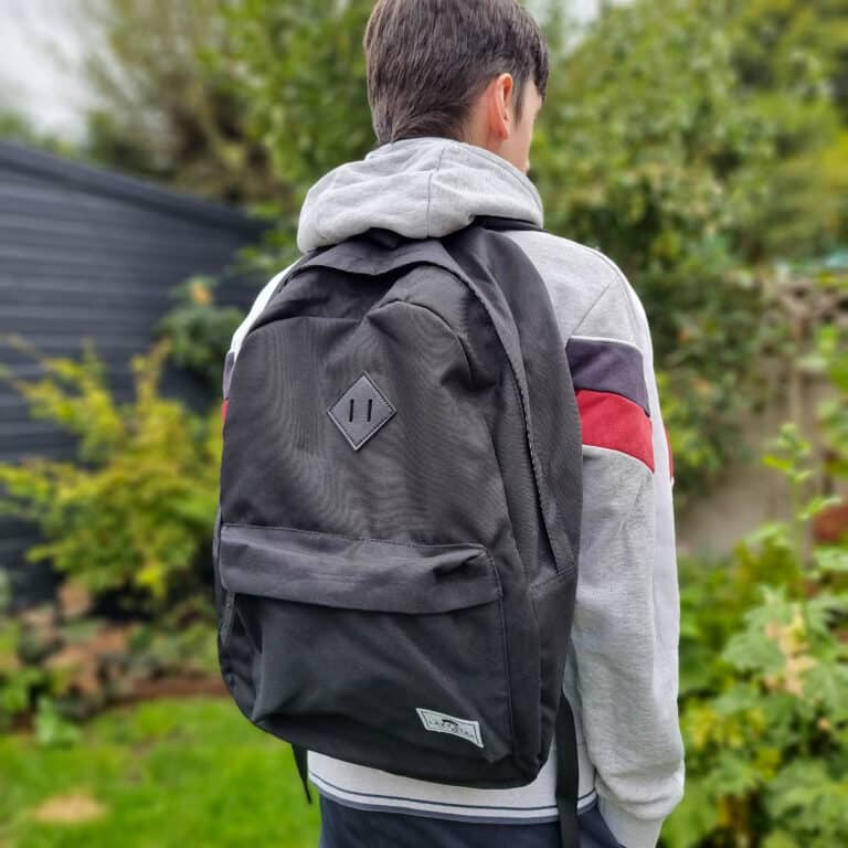 A person wearing a grey hoodie with red and navy stripes stands outdoors, facing away, with a large black rucksack—perhaps taking a break from their work from home environment. Greenery and a dark fence are visible in the background.