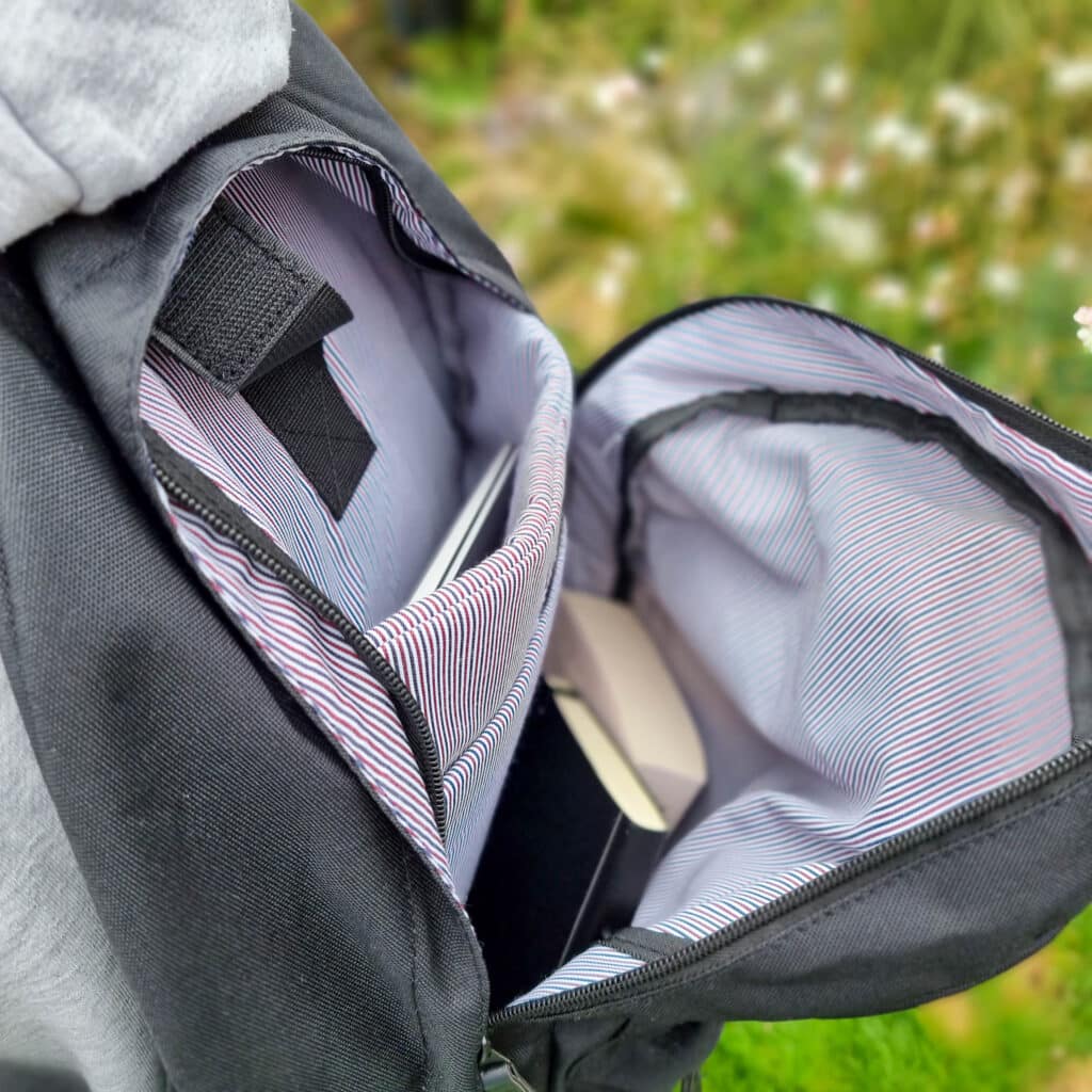 An open black rucksack with a striped inner lining, showing a book and a closed laptop inside—ideal essentials to create a good work from home environment. The background is grassy and slightly blurred.