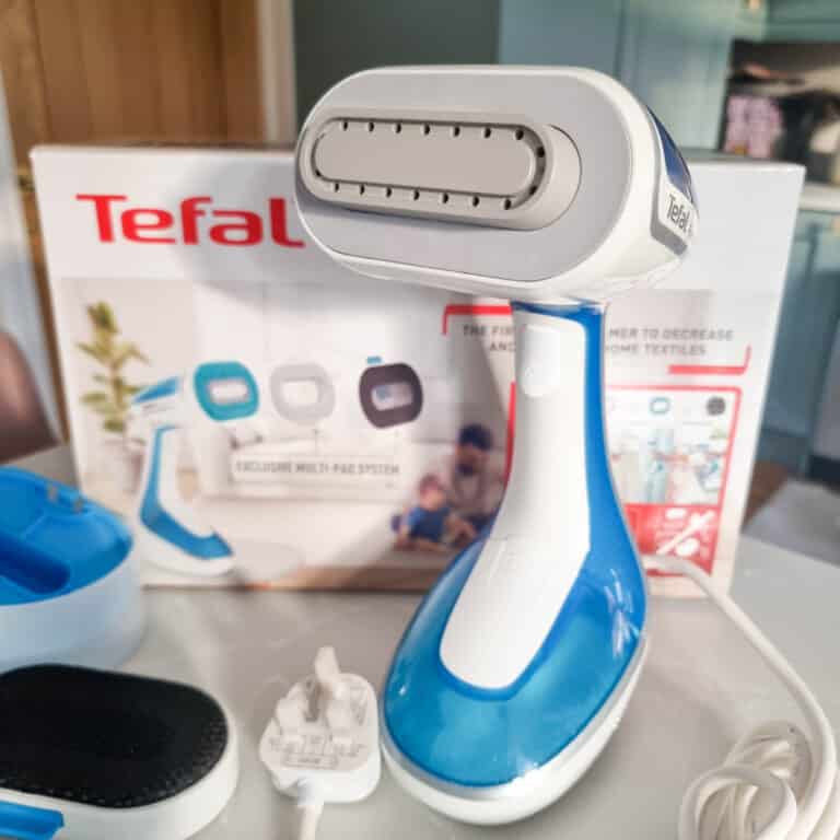 A blue and white Tefal DT9530 Handheld Garment Steamer sits on a table with its box and accessories, including a plug adaptor and brush attachment, visible in the background. Perfect for those seeking an honest review of its features.
