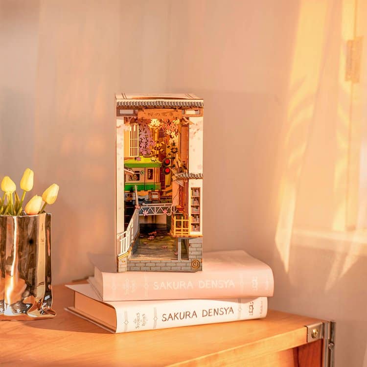 A small, detailed diorama of a cosy room sits on two stacked books titled SAKURA DENSYA, next to a metallic vase holding yellow tulips—like a stylish puzzle, all are bathed in warm sunlight.