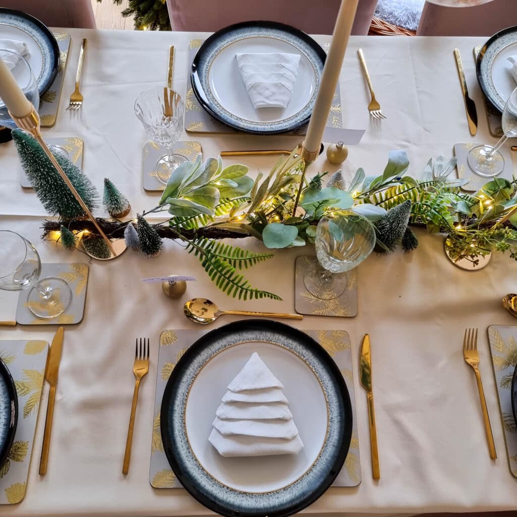A Green Christmas table features gold cutlery, black-rimmed plates, and napkins folded into Christmas tree shapes. The centrepiece displays greenery, small trees, fairy lights, and gold baubles on a cream tablecloth.
