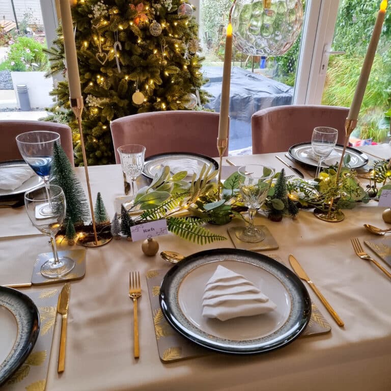 A festive Green Christmas table is set with gold cutlery, plates, serviettes folded like Christmas trees, glasses, and green, gold, and white decorations. A decorated Christmas tree with lights glows in the background near a window.