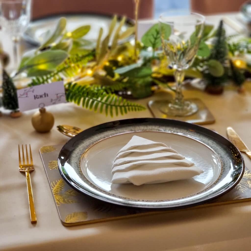 Elegant Green Christmas table setting with a white serviette folded into a tree shape on a plate, surrounded by gold cutlery and glasses. Greenery and fairy lights create a festive centrepiece, with a place card reading Ruth.