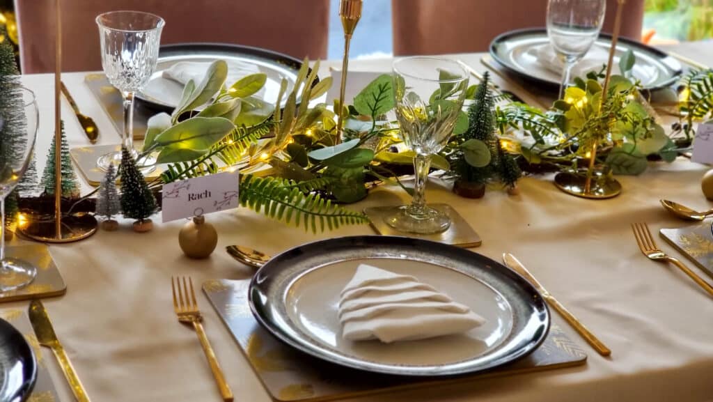 Create a stunning Gold Christmas Table with gold cutlery, white plates, and serviettes folded like trees. Greenery, fairy lights, and small pine tree decorations adorn the cream-coloured tablecloth for a magical holiday setting.