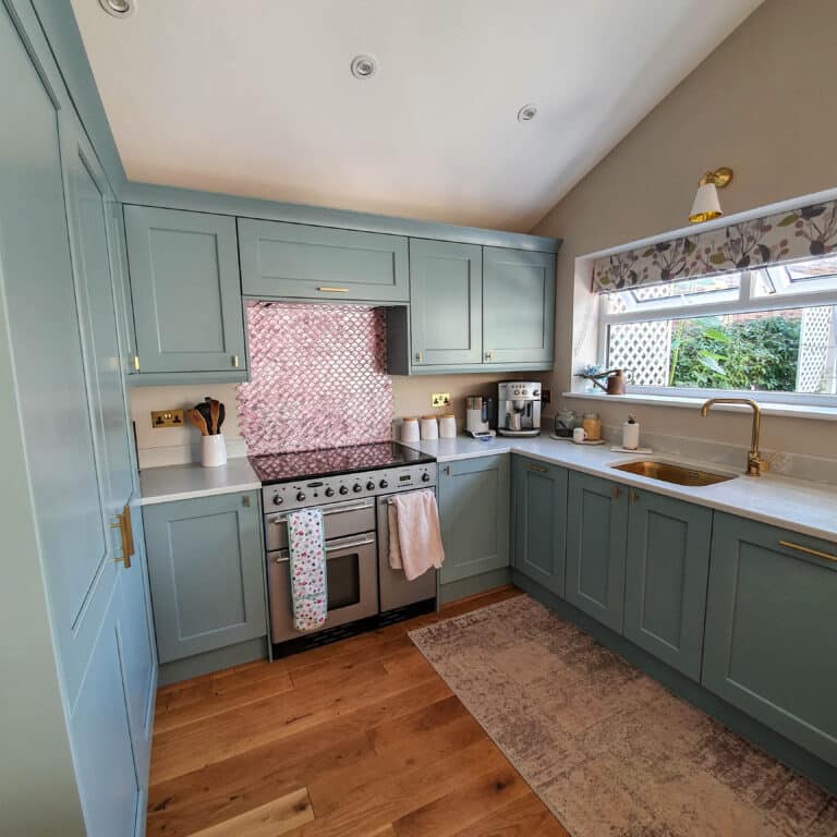 A modern kitchen with light blue kitchen cabinetry, a stainless steel oven, a pink tile splashback, wooden floor, gold fittings, and a window above the sink letting in natural light—perfect for easy cupboard maintenance.