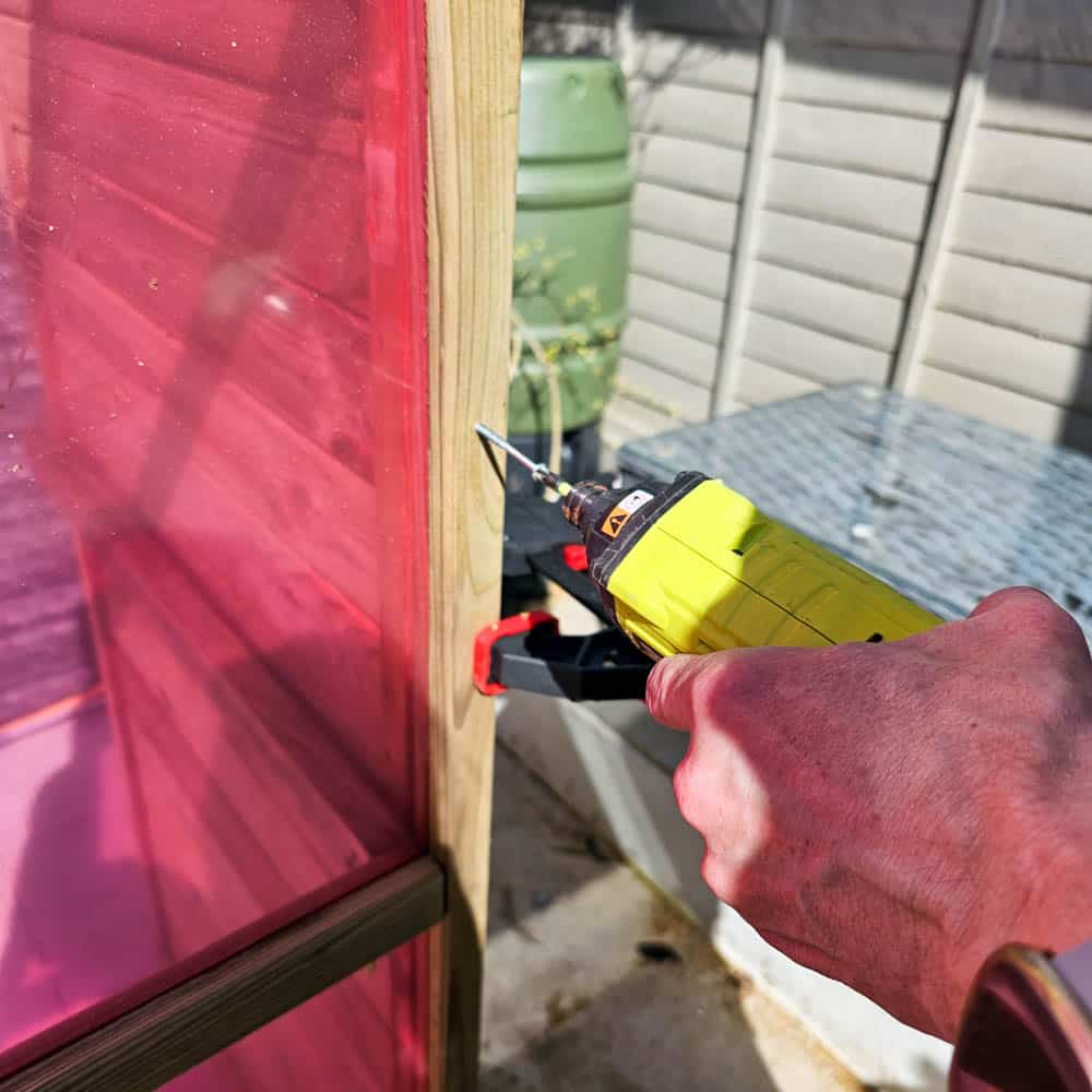 A person uses a yellow staple gun to secure pink plastic sheeting to a wooden frame outdoors, near a Wooden Greenhouse and garden area, perfect for a quick Greenhouse Review.