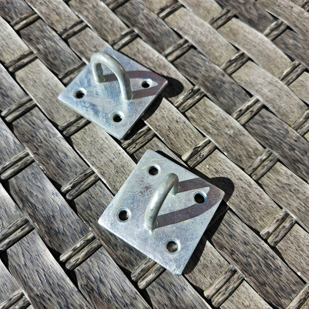 Two metal mounting plates with curved hooks and four screw holes each, displayed on a weathered wooden surface in bright sunlight—perfect hardware for a wooden greenhouse or complementing a Victorian wooden greenhouse design.