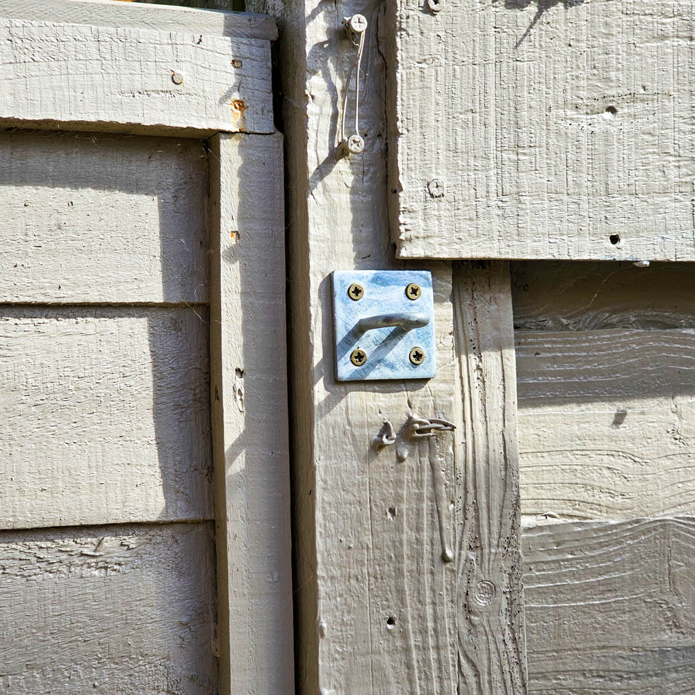 Close-up of a weathered wooden fence with a small, silver metal bracket attached by six screws. The light grey wood shows texture, knots, and wear—reminiscent of materials found in a classic Forest Victorian Greenhouse.