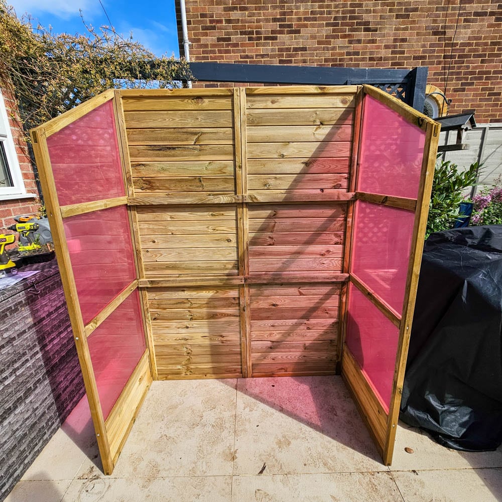 A 5ft Wooden Greenhouse with three sides and clear panels tinted pink on the left and right stands on a patio next to a brick wall, with garden plants in the background.