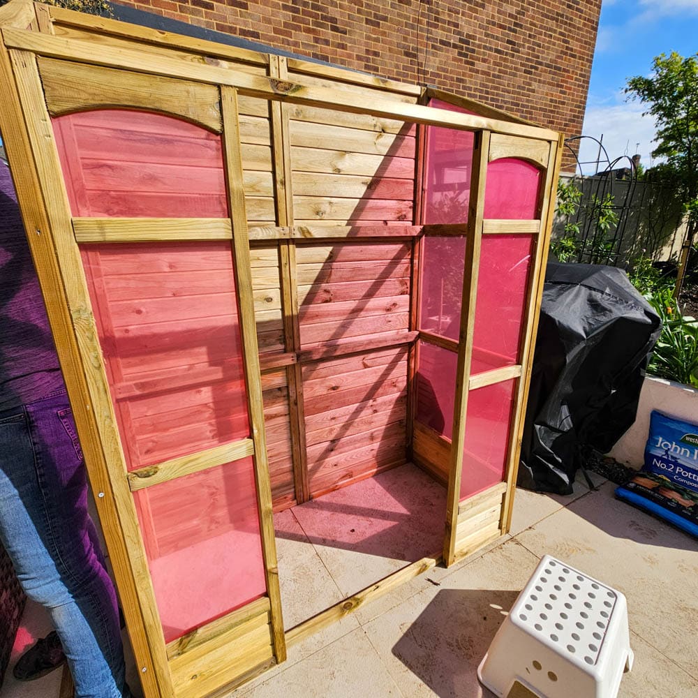 A partially assembled Forest Victorian Wooden Greenhouse frame with pink translucent panels stands outdoors on a patio near a brick wall and some garden items. A person stands to the left, mostly out of view.