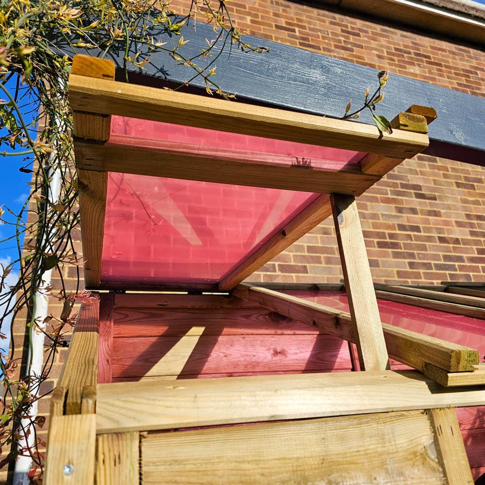 A close-up, upward view of a wooden greenhouse structure with angled red translucent panels, set against a brick wall and partly shaded by climbing plant stems under a blue sky.