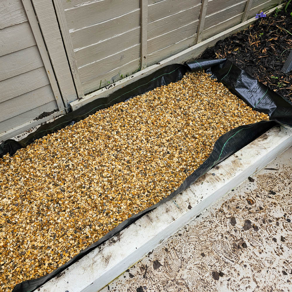 A rectangular garden bed lined with black fabric and filled with small golden gravel sits next to a wooden fence, a 5ft Wooden Greenhouse, and a concrete path, with soil and mulch visible at one end.