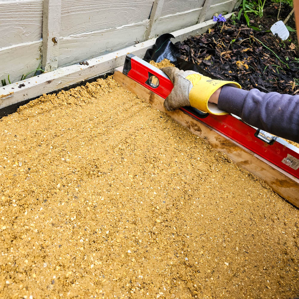 A person wearing a glove uses a red spirit level to smooth and flatten compacted sand or gravel in a garden bed beside a white wooden fence, preparing the area for a 5ft greenhouse or Forest Victorian greenhouse.