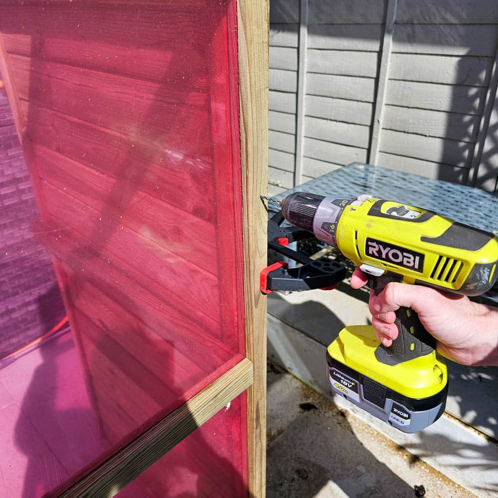 A person uses a yellow Ryobi cordless drill to screw a wooden greenhouse frame with pink translucent panels outdoors, next to a grey fence and a blue metal table—perfect for any Victorian greenhouse or greenhouse review.