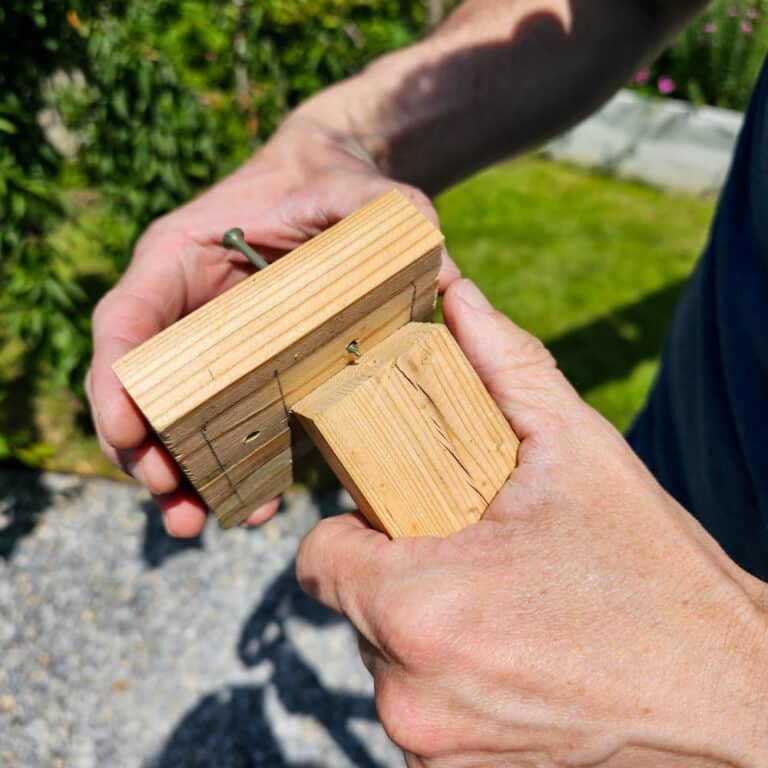 A person holds two pieces of wood joined at a right angle, fastening them together with a screw outdoors on a sunny day, demonstrating how to build a wooden obelisk step-by-step.