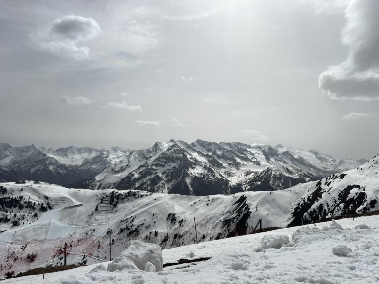 Snow-covered mountain range under a cloudy sky with scattered light, featuring a ski piste and red safety netting in the foreground—Austria Unveiled in its winter splendour.