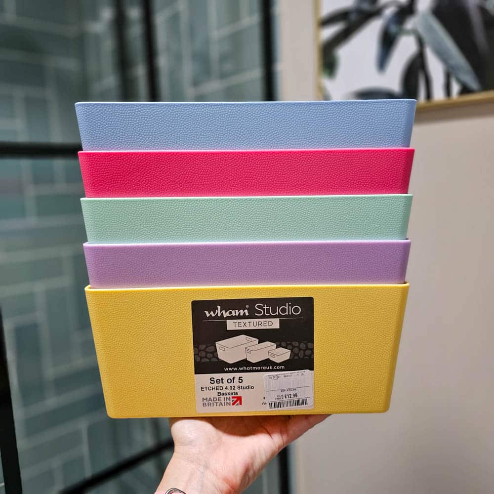 A hand holds a set of five rectangular textured storage baskets in yellow, lavender, mint green, pink, and blue—perfect for organising during renovations. The baskets are stacked and labelled “wham Studio,” with a blurred indoor background visible.