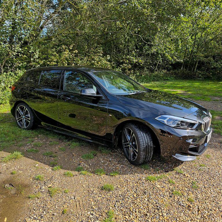 A shiny black BMW hatchback is parked on a gravel area beside lush grass, surrounded by green trees and bushes at Sarah Raven Gardens—a perfect Garden Getaway on a sunny day.