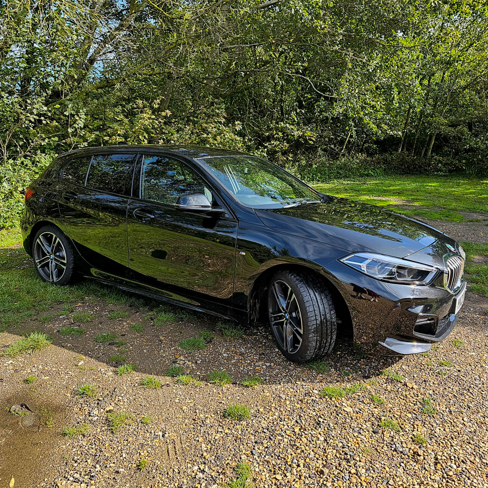 A shiny black BMW hatchback is parked on a gravel area beside lush grass, surrounded by green trees and bushes at Sarah Raven Gardens—a perfect Garden Getaway on a sunny day.