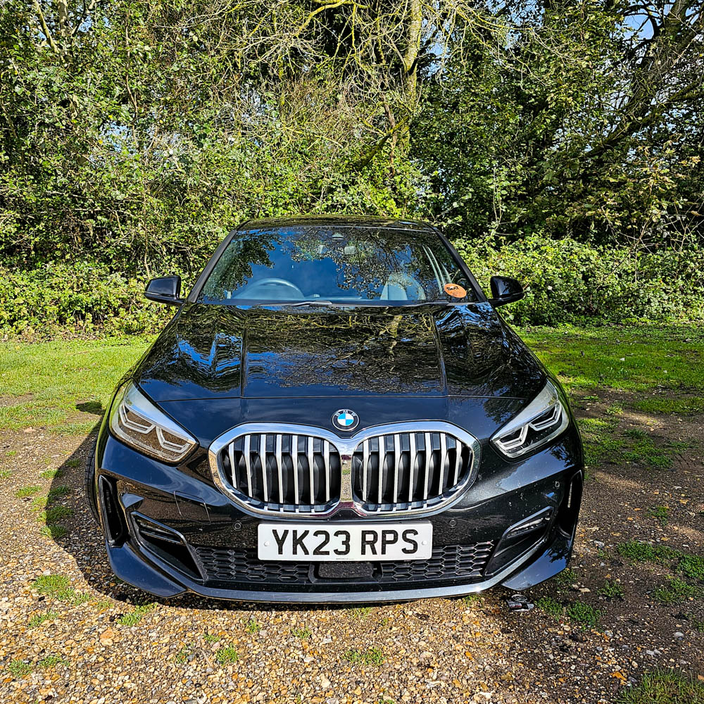 A black BMW with number plate YK23 RPS is parked on a gravel path surrounded by lush greenery—perfect for a Garden Getaway near Sarah Raven Gardens.