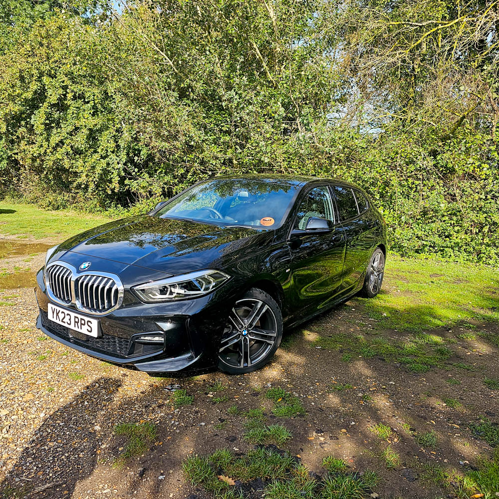 A black BMW hatchback is parked on a grassy area near trees and bushes at Sarah Raven Gardens, with sunlight reflecting off its bonnet. The car has the registration plate YK23 RPS—a perfect Garden Getaway found via Discovercars.