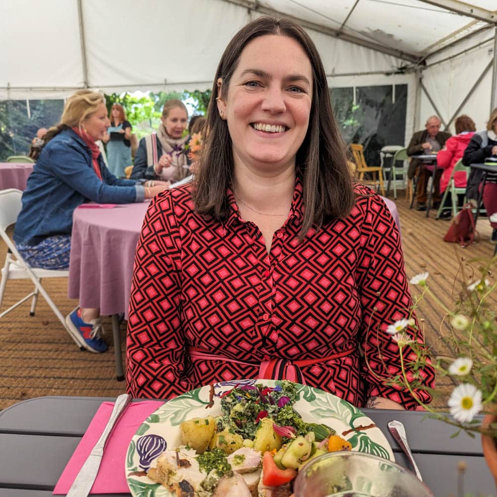 A woman with long brown hair, wearing a red patterned dress, sits smiling at a table with a plate of food in front of her inside a white marquee at Sarah Raven Gardens, with other people dining in the background.