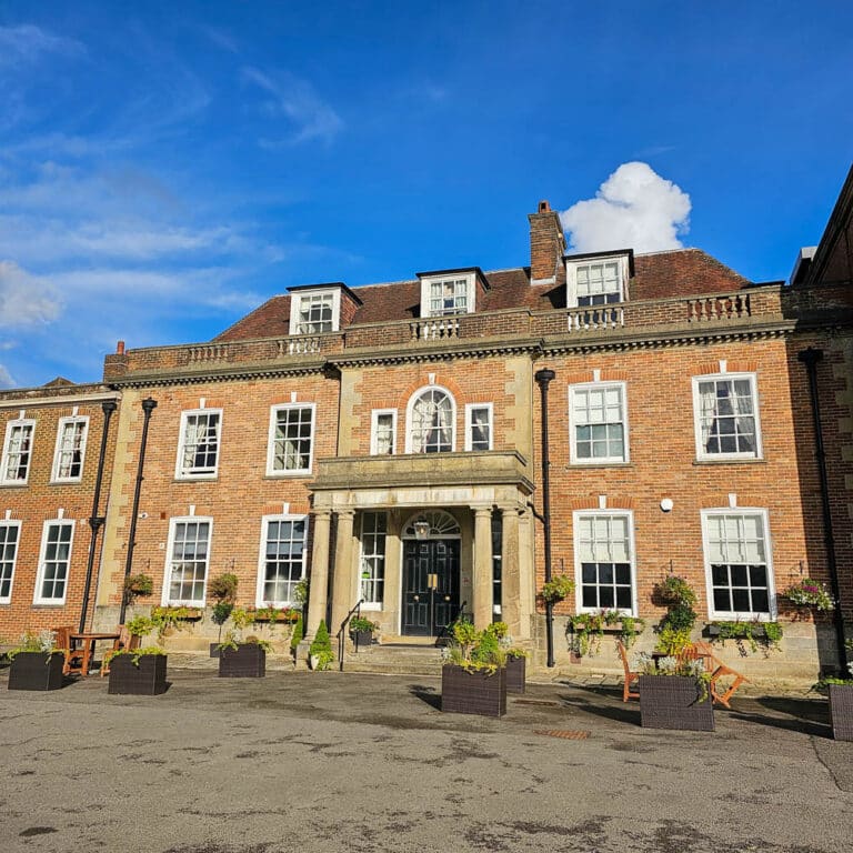 A large, historic brick building with white-trimmed windows and a central arched entrance, featuring a stone porch with columns and planters—an ideal dream setting for an unforgettable engagement venue under a clear blue sky.