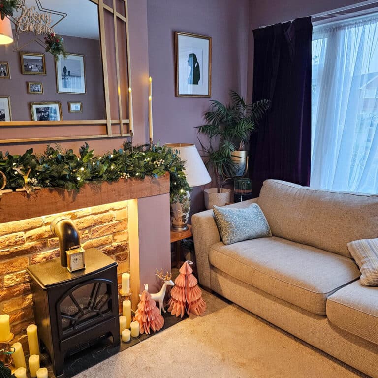 A cosy living room with a beige sofa, decorated fireplace, pink Christmas tree baubles, framed pictures, a potted plant, and sheer curtains letting in natural light—perfect for relaxing after reading an Air Purifier Review or a Briiv Air Purifier One Year Update.
